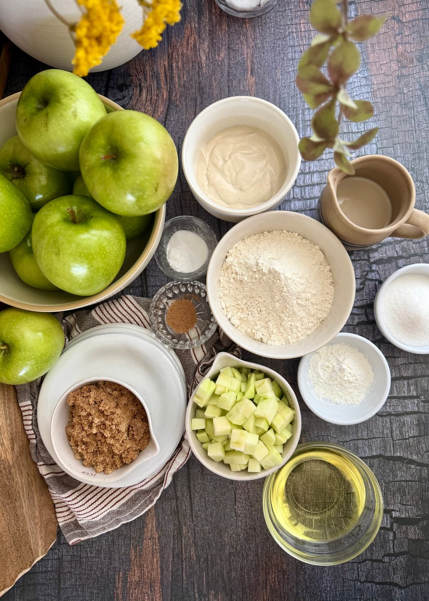 Apple cake ingredients including chopped apples, yogurt, milk, flour, sugars, and oil arranged on a table