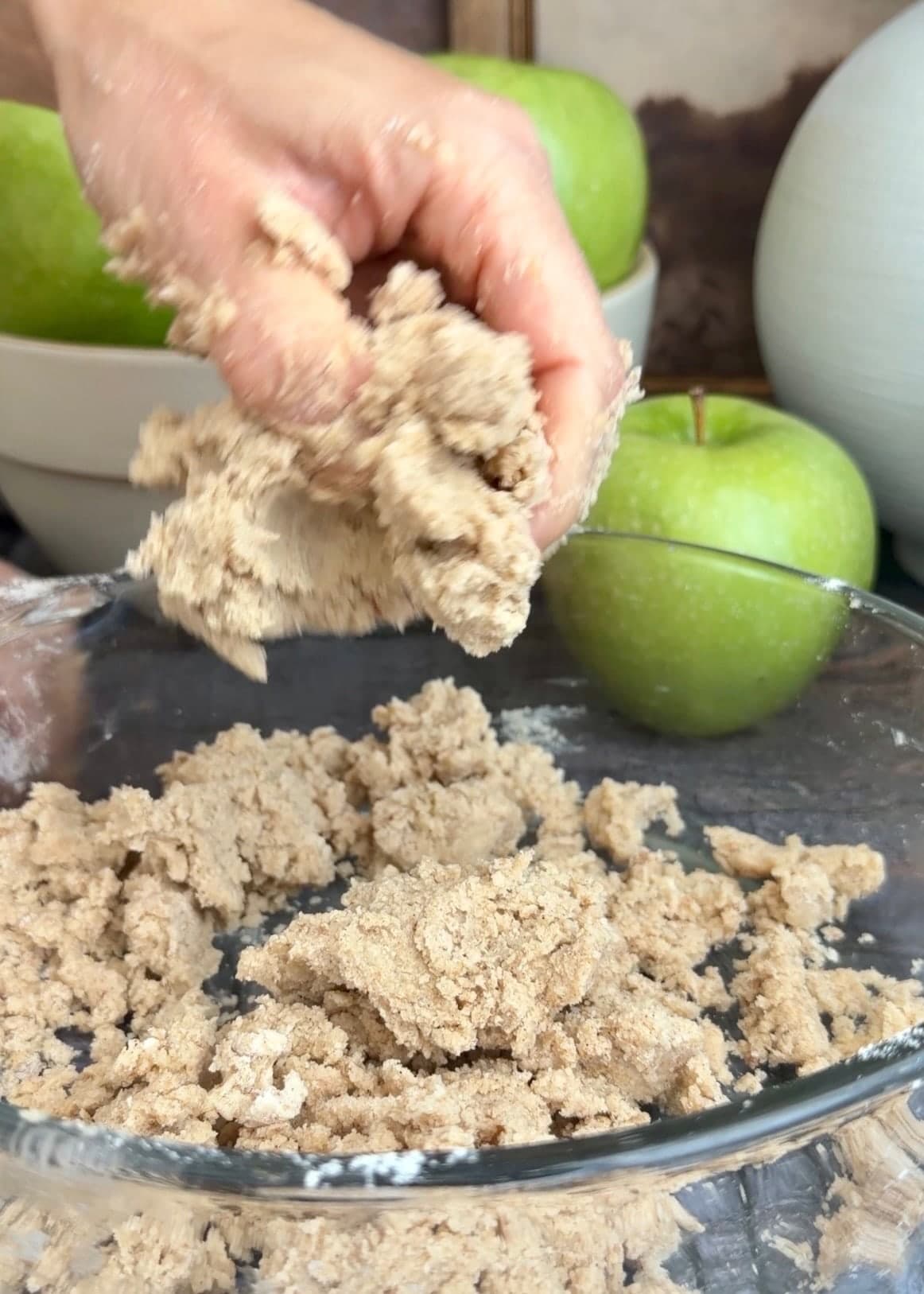 A hand squeezing large pieces of crumble over a glass bowl