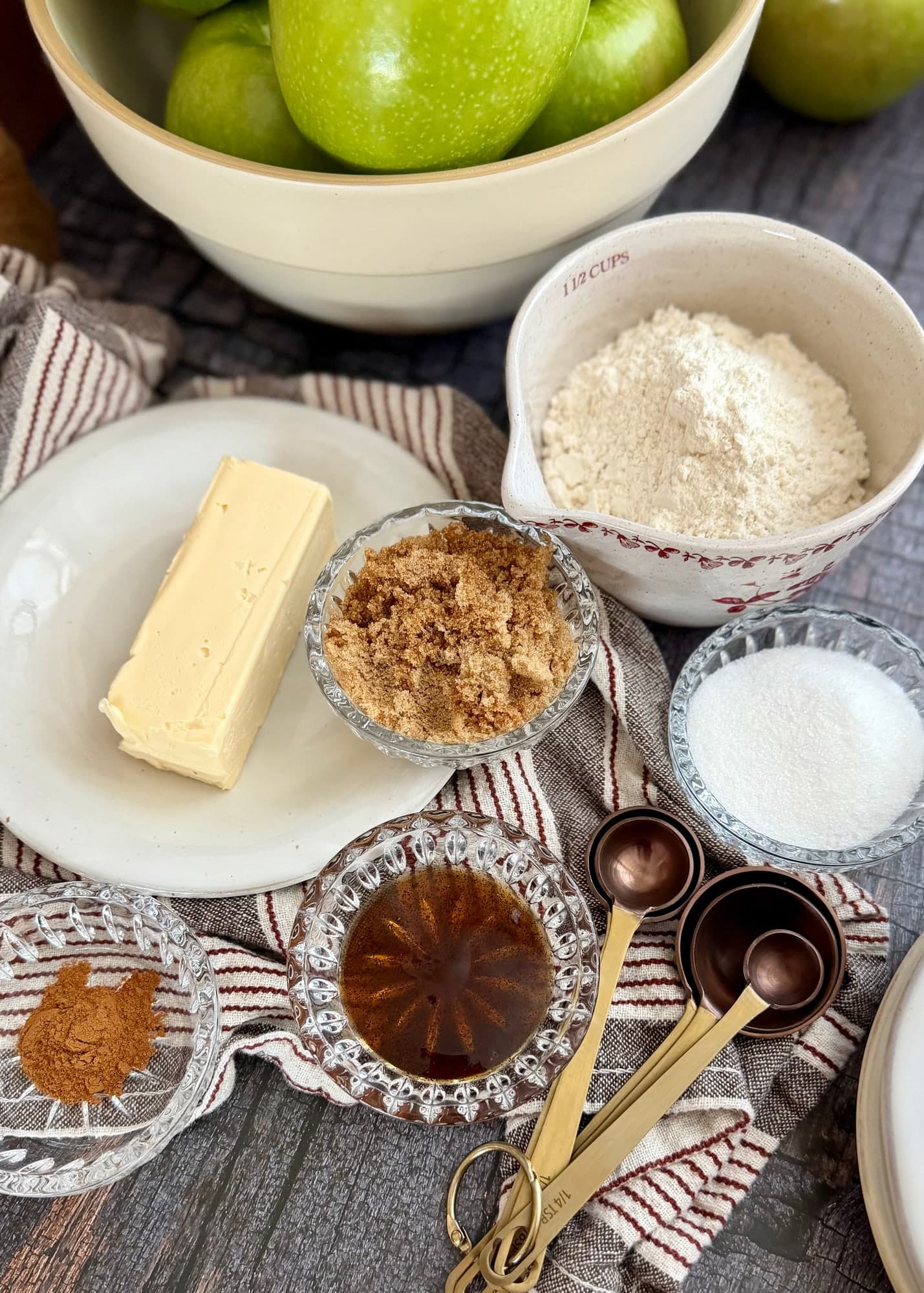 Butter, flour, brown sugar, granulated sugar, cinnamon, and vanilla arranged beside green apples