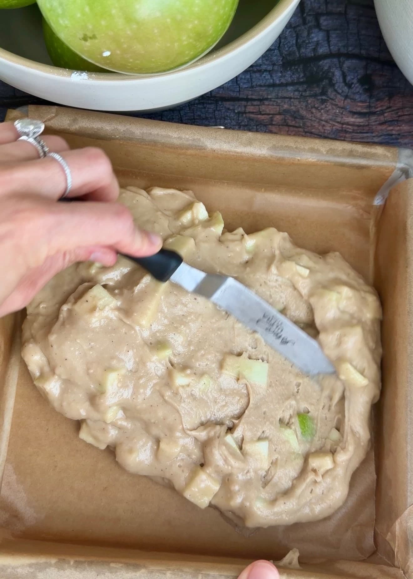 An offset spatula spreading apple batter inside a parchment-lined square pan