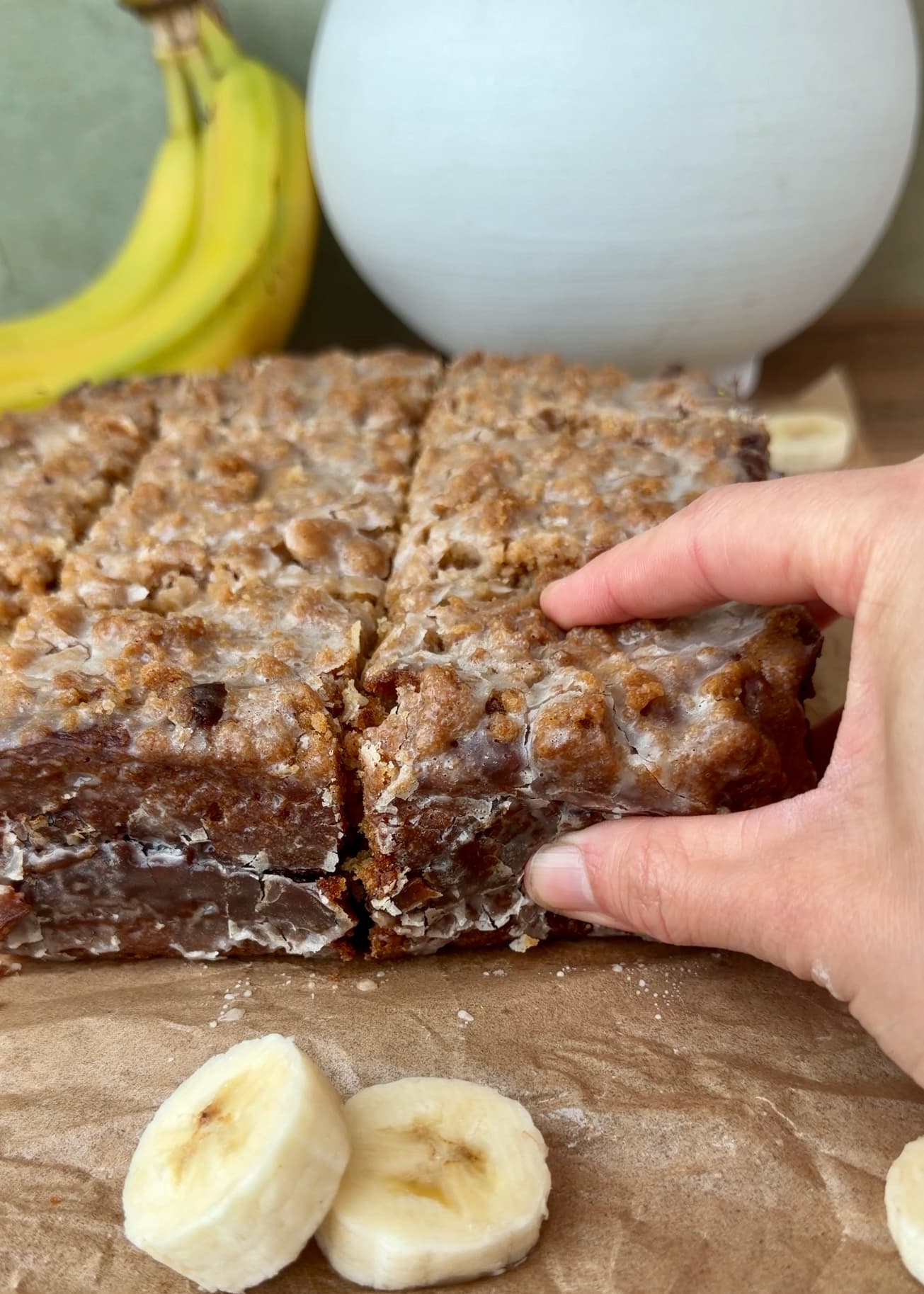 A hand lifting a square from the glazed banana coffee cake