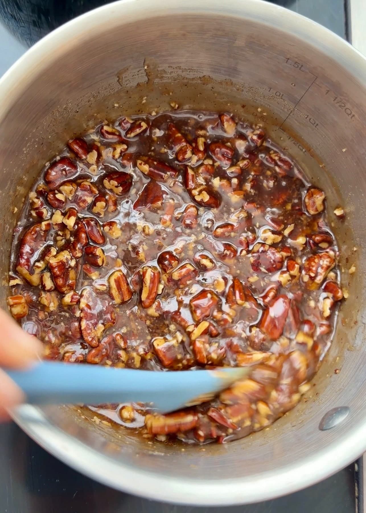 A blue spatula stirring chopped pecans through the glossy topping