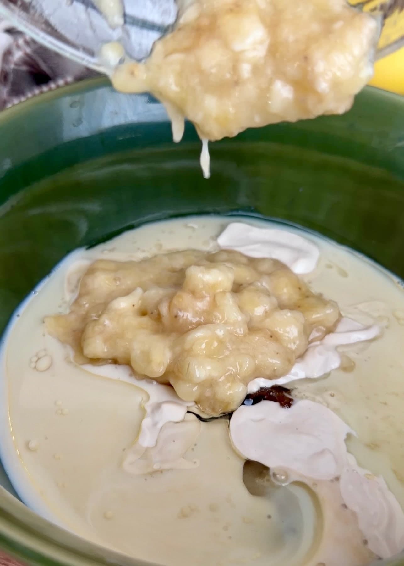 Mashed banana being added to the wet ingredients in a green mixing bowl
