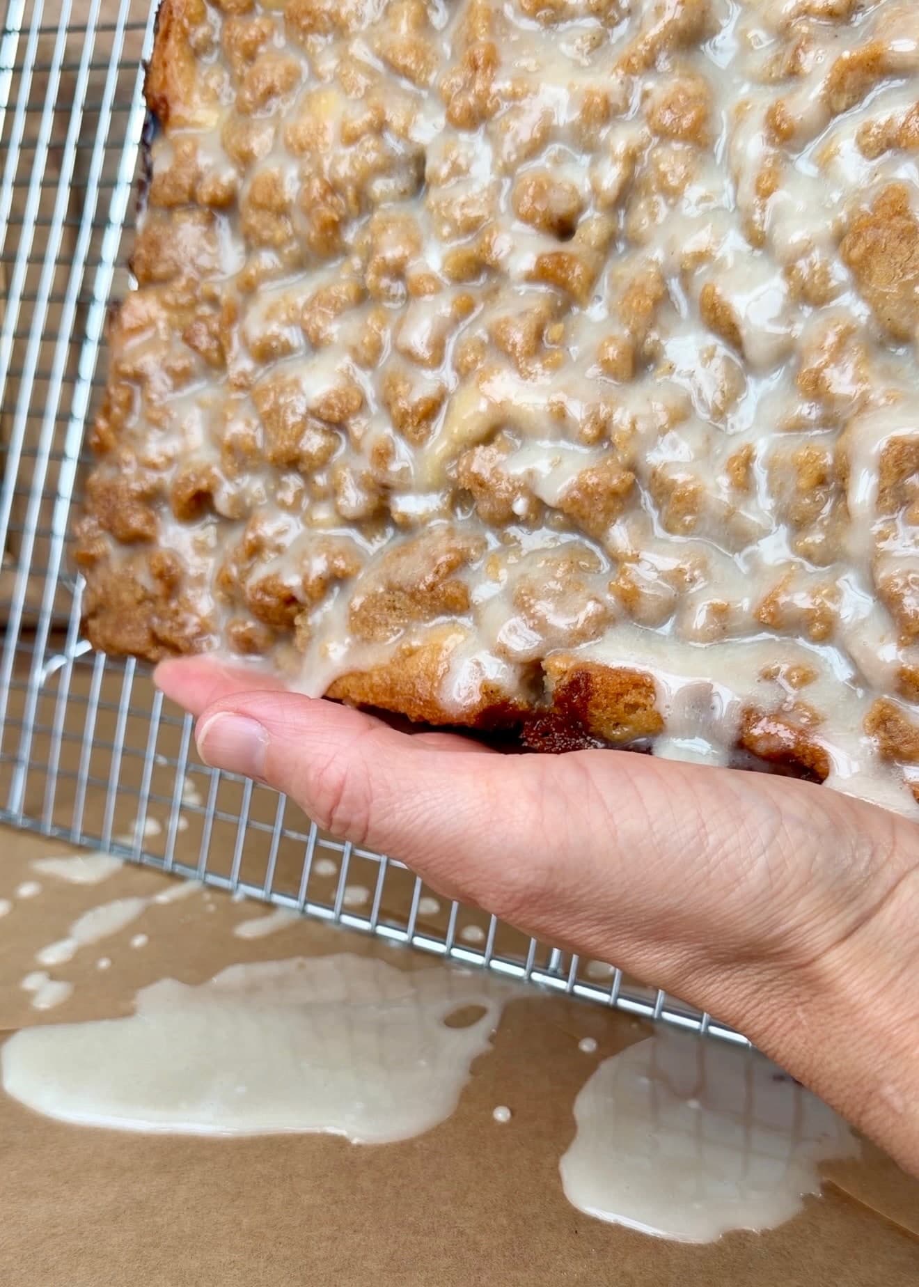 A hand supporting the tilted glazed cake as excess icing collects below the rack