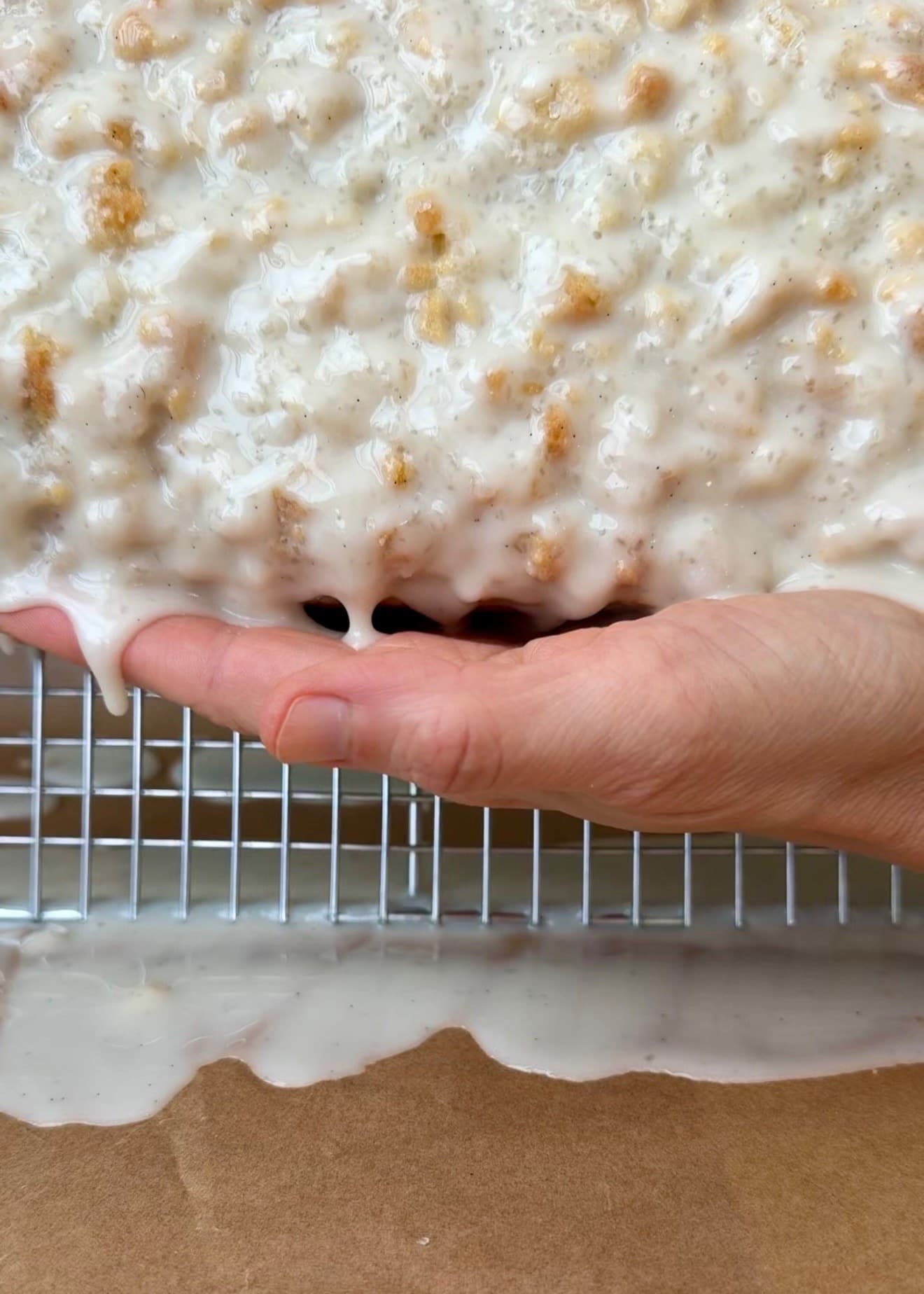A hand supporting the glazed loaf as it is tilted above the rack, with glaze dripping toward the tray