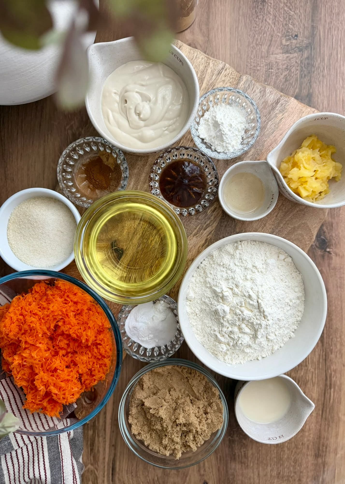 Carrots, pineapple, flour, sugars, yogurt, and other carrot cake ingredients arranged on a table