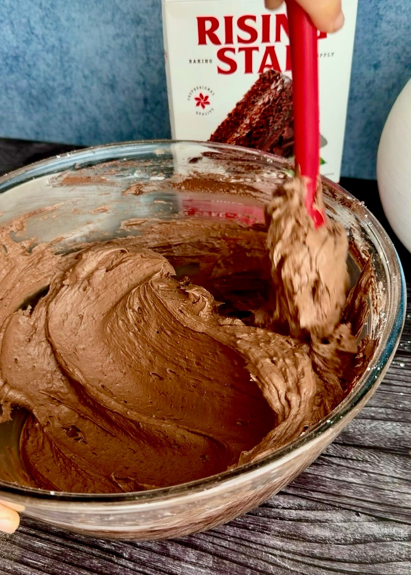 A red spatula smoothing chocolate frosting against the side of the bowl