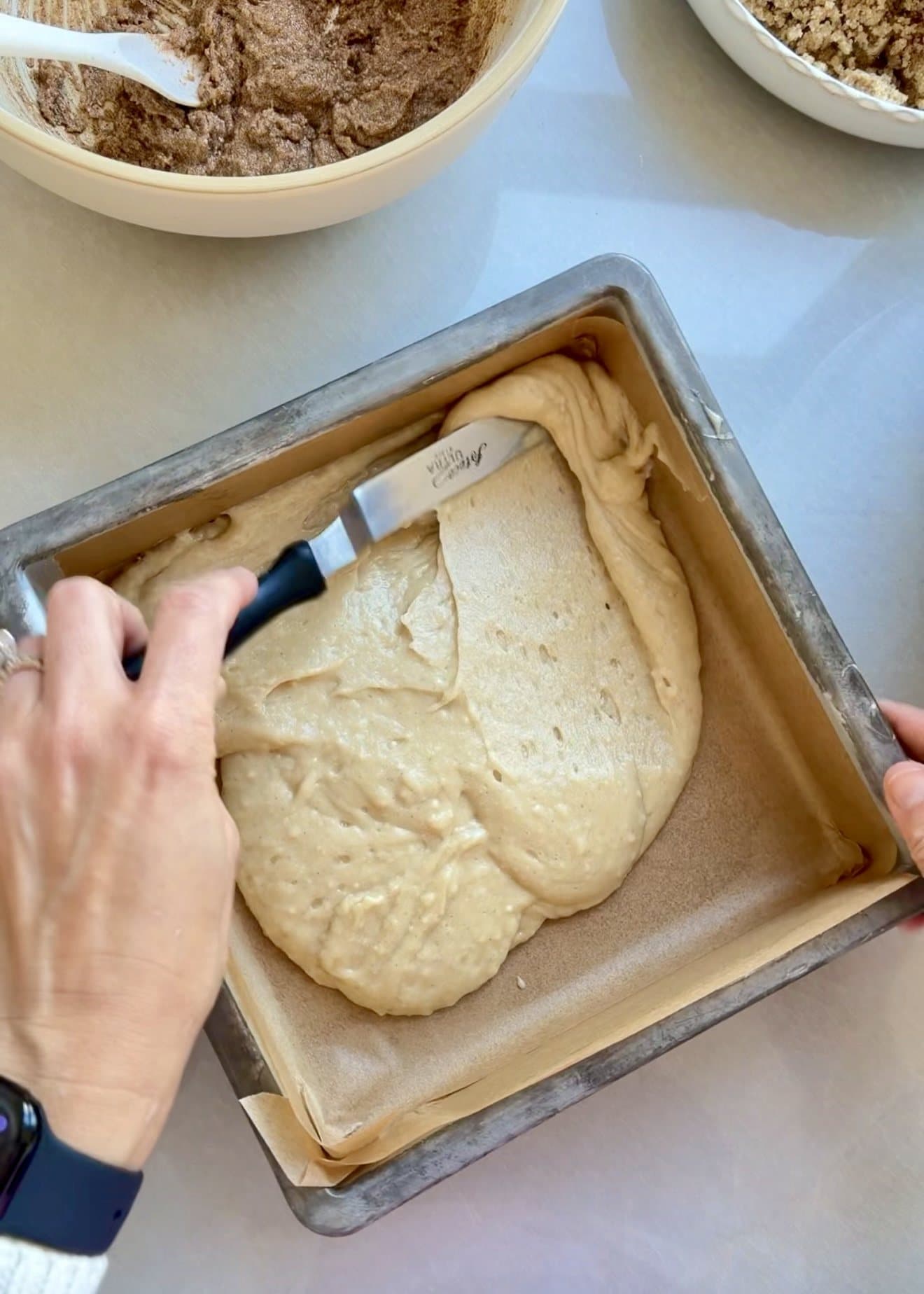 First half of the vanilla coffee cake batter being spread in a parchment-lined 9-inch square pan