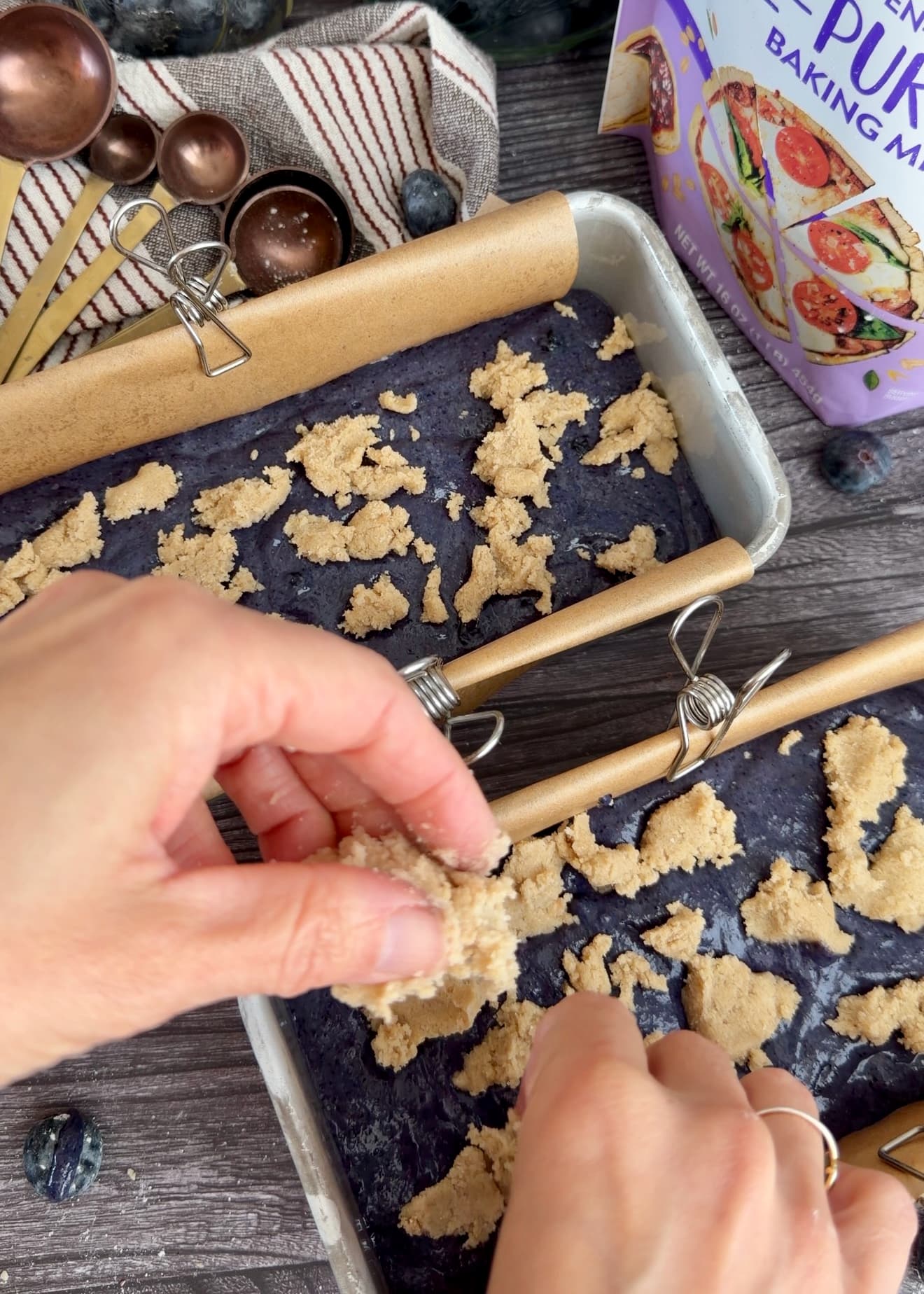 Hands scattering pale crumble over blueberry batter in mini loaf pans