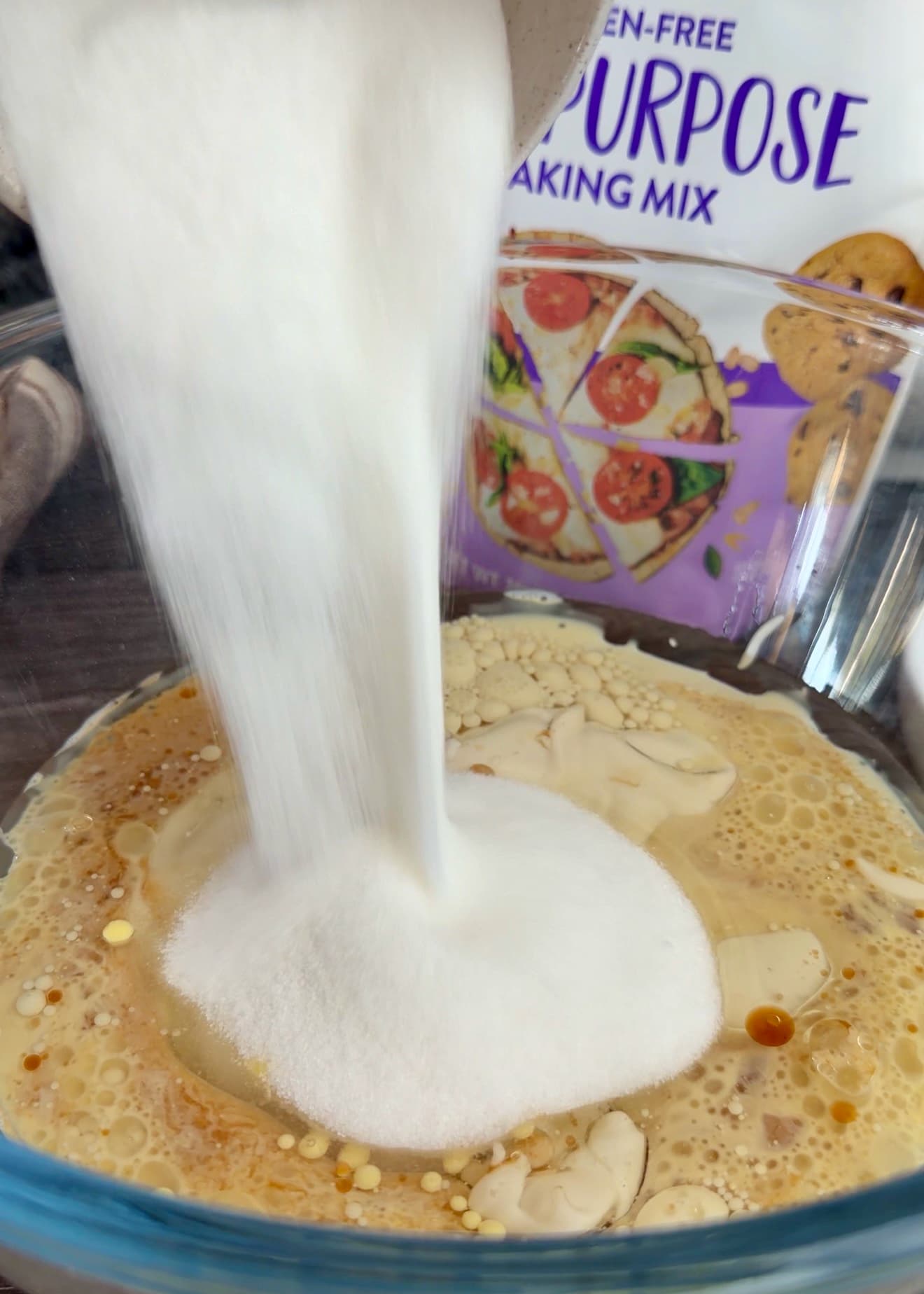 Sugar being poured into the wet ingredients in a glass bowl