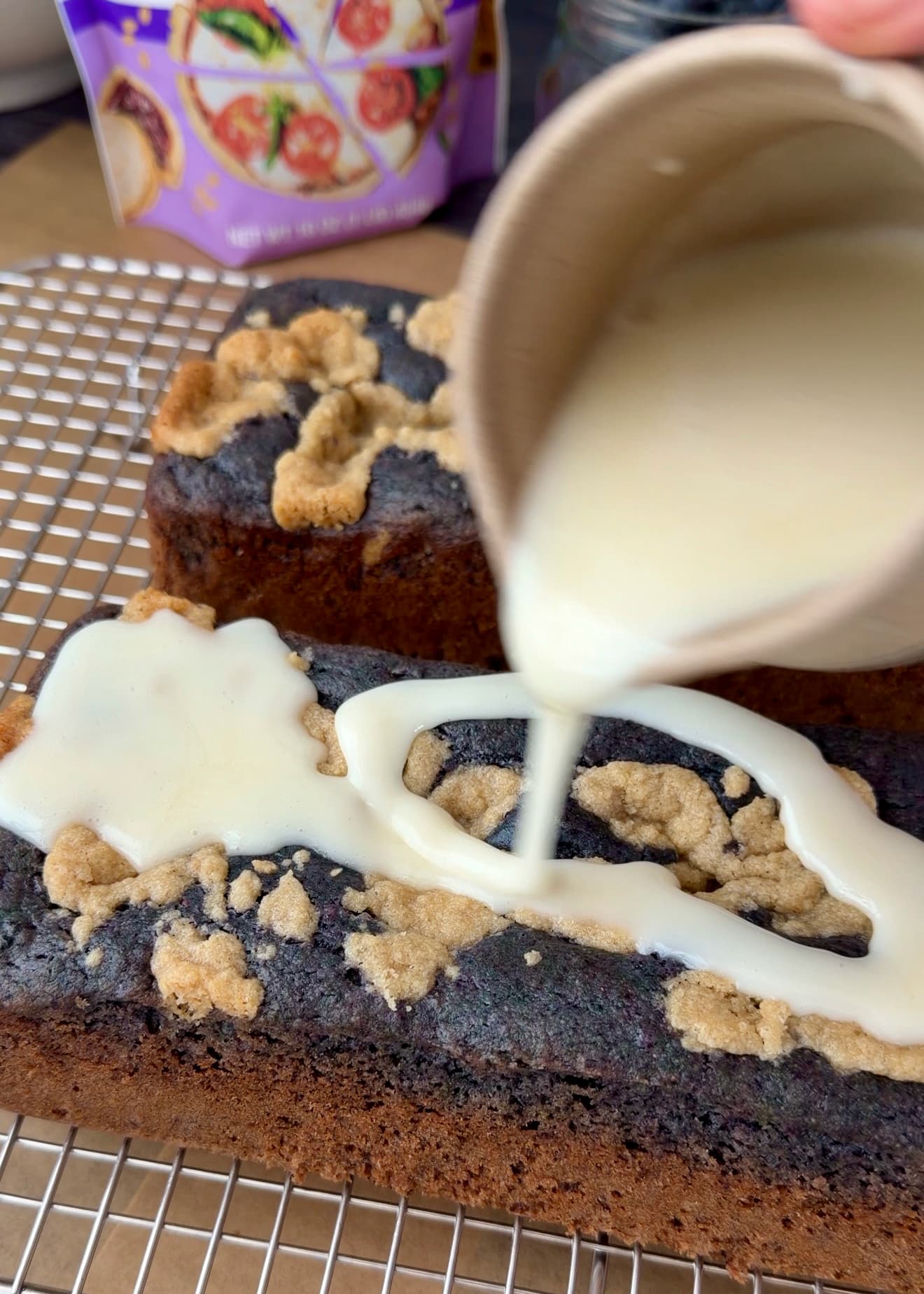 Pale glaze pouring from a pitcher over a blueberry loaf on a wire rack