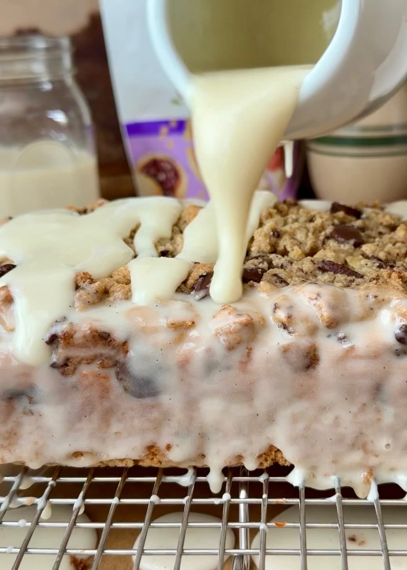 Vanilla glaze pouring from a white pitcher over the streusel loaf on a rack