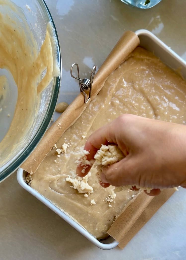 A hand scattering cold streusel over the first layer of loaf batter
