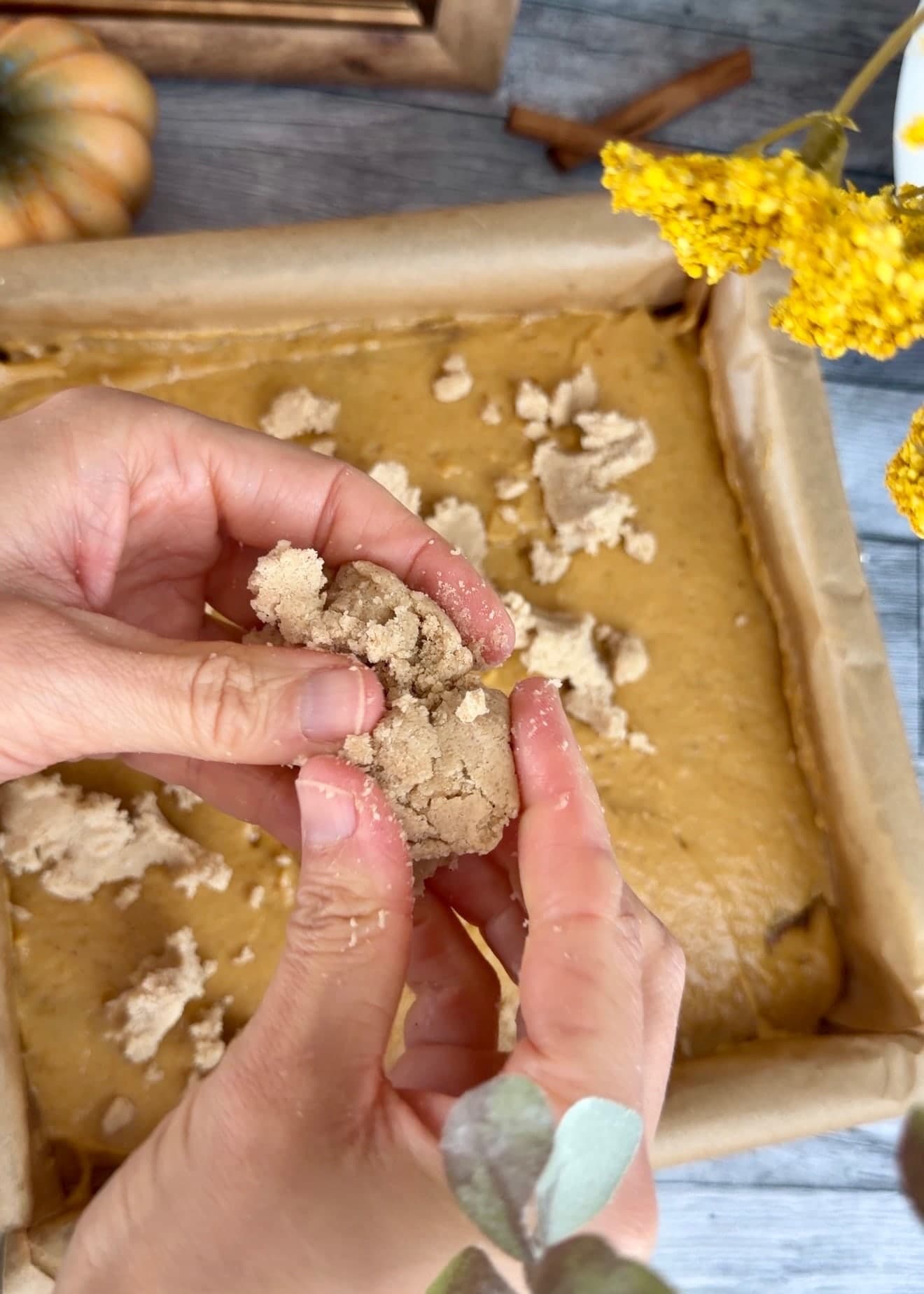 Hands breaking chilled crumble into pieces over the pumpkin batter