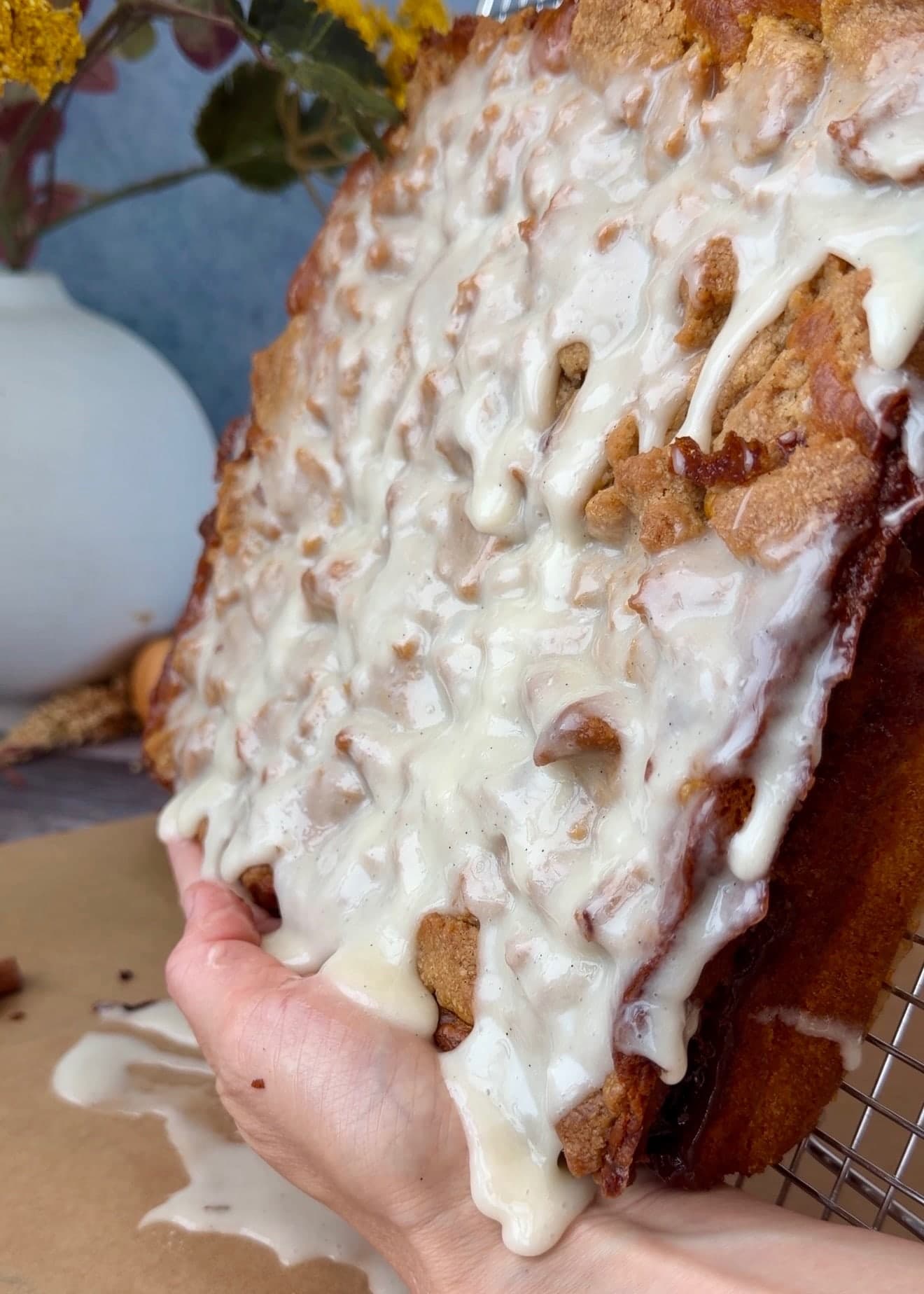 Hands holding the cake at an angle as excess glaze drips onto the tray below