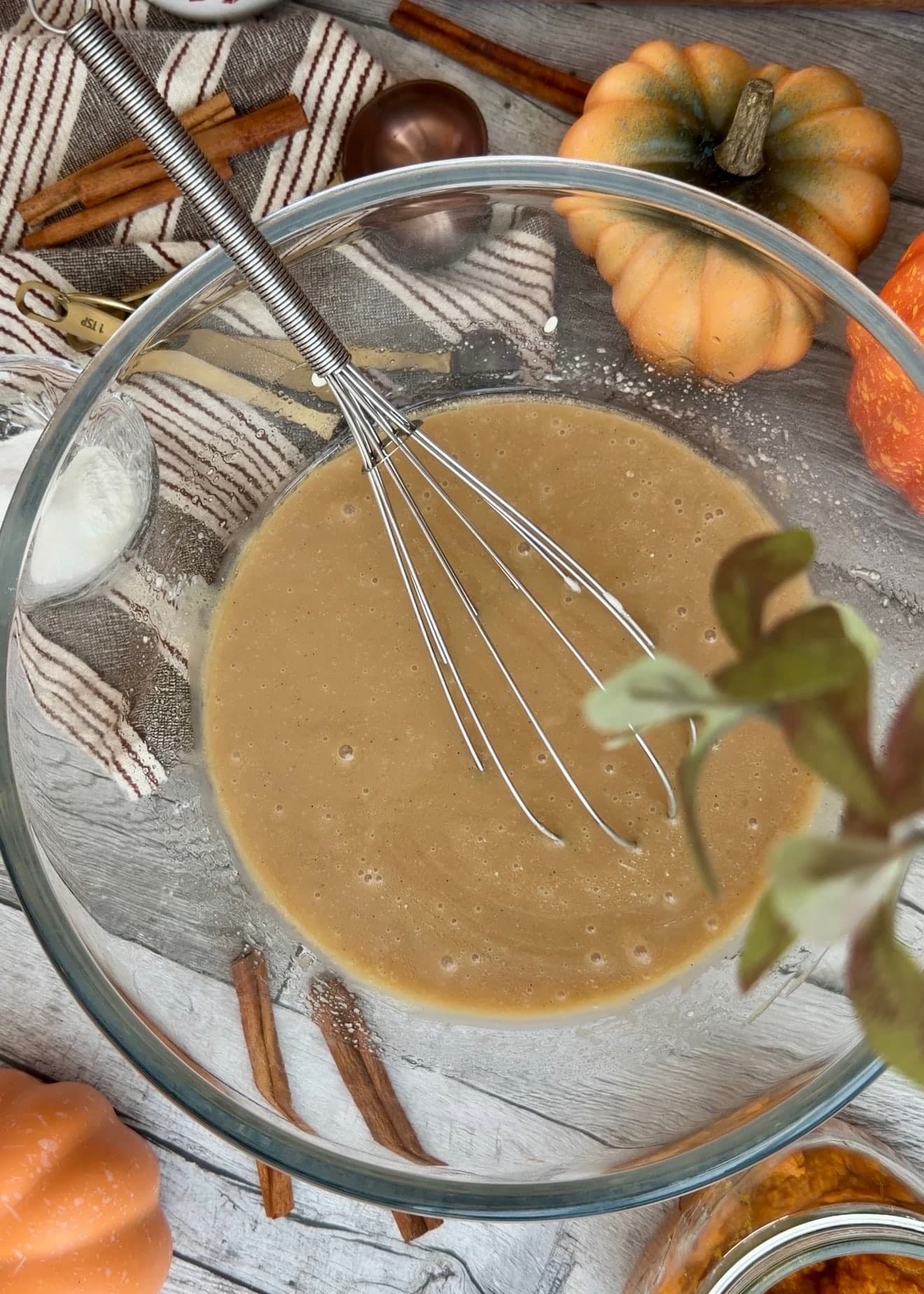 A whisk resting in a smooth tan mixture in a glass bowl