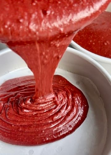 Red velvet batter pouring into a lined round cake pan