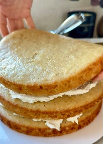 Hands lowering a third vanilla cake layer onto a frosted stack