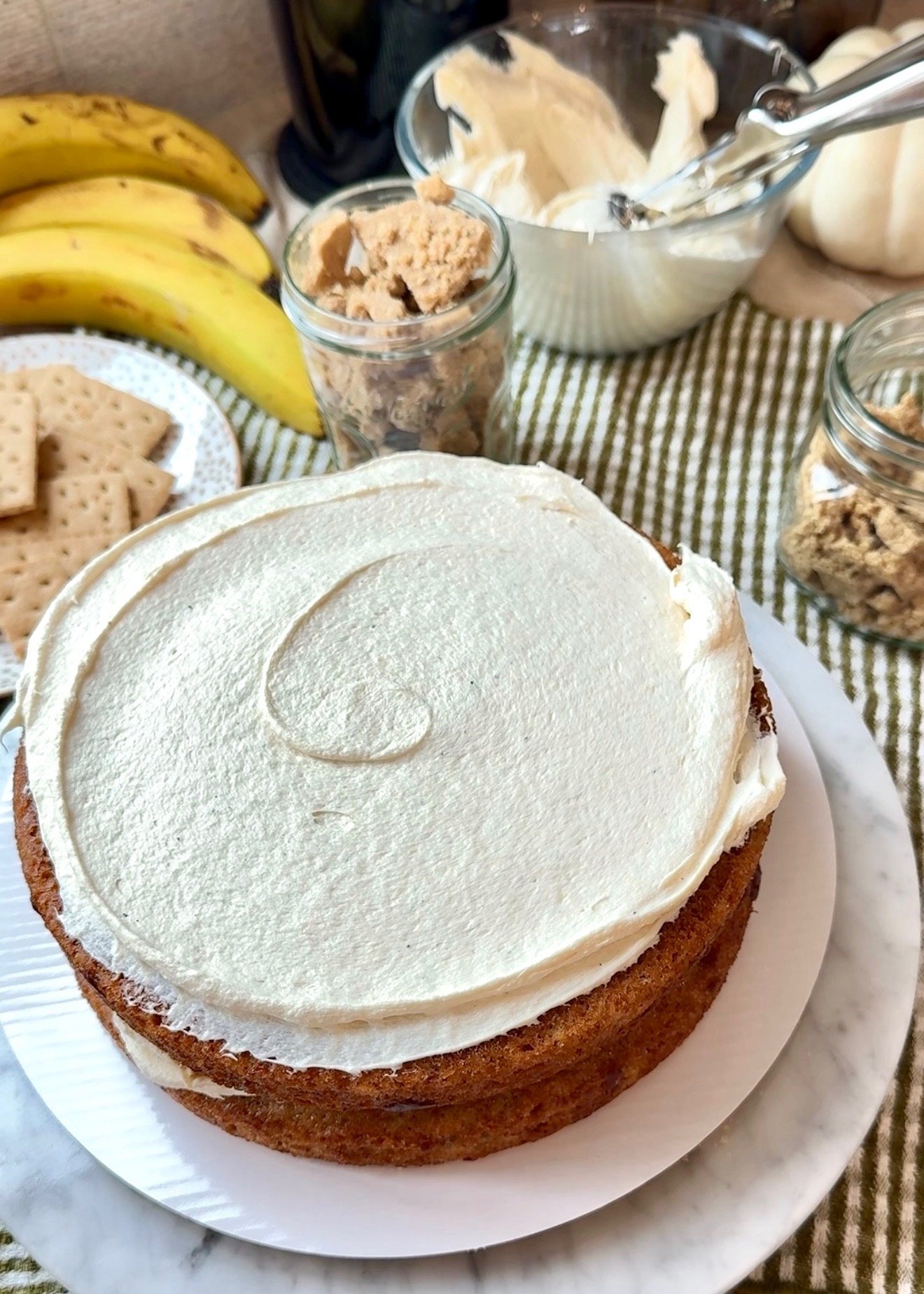 Velvety cheesecake frosting spread over warm banana cake with bananas and graham crackers nearby